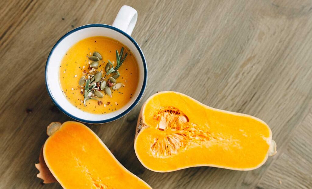 White mug of butternut squash with seeds and seasonings on top. Also with a butternut squash cut in half next to it.