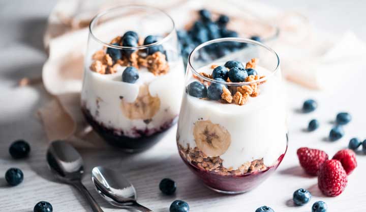 Two dessert glasses with coconut cream, berries, bananas, and nuts.