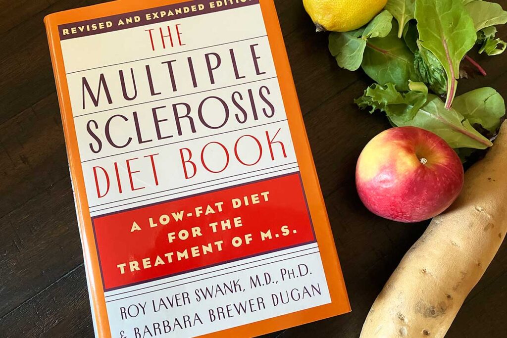 The Multiple Sclerosis Diet Book on a dark table with veggies.