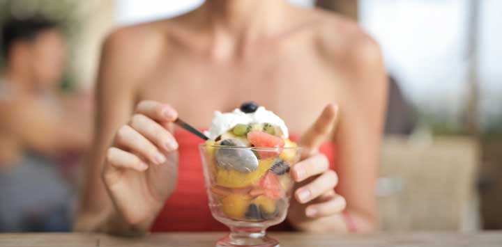 Woman eat a glass bowl of fruit topped with coconut cream.