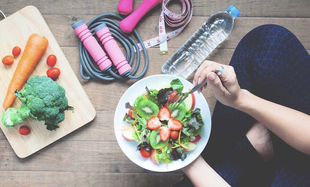 Woman sitting on floor eating a salad with jump rope, weights, and a water bottle.