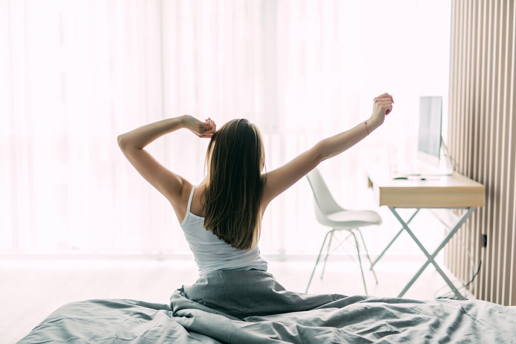 Back view of young woman stretching on unmade bed after waking up.