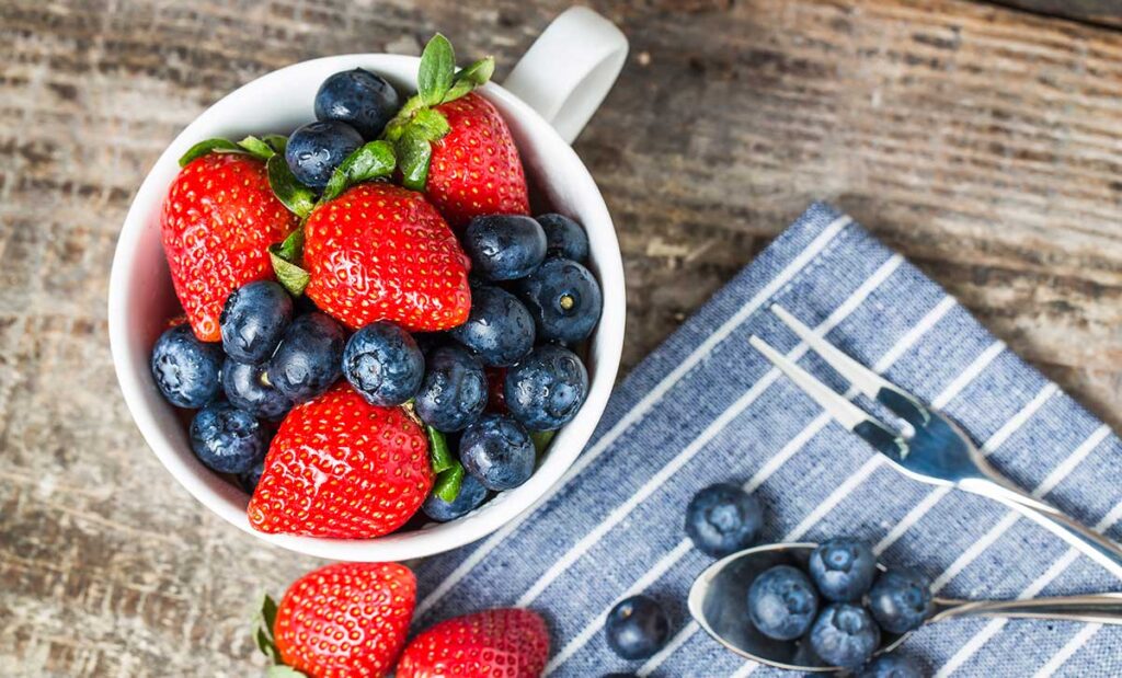 Blueberries and strawberries in a white mug with some on a blue and white striped napkin.