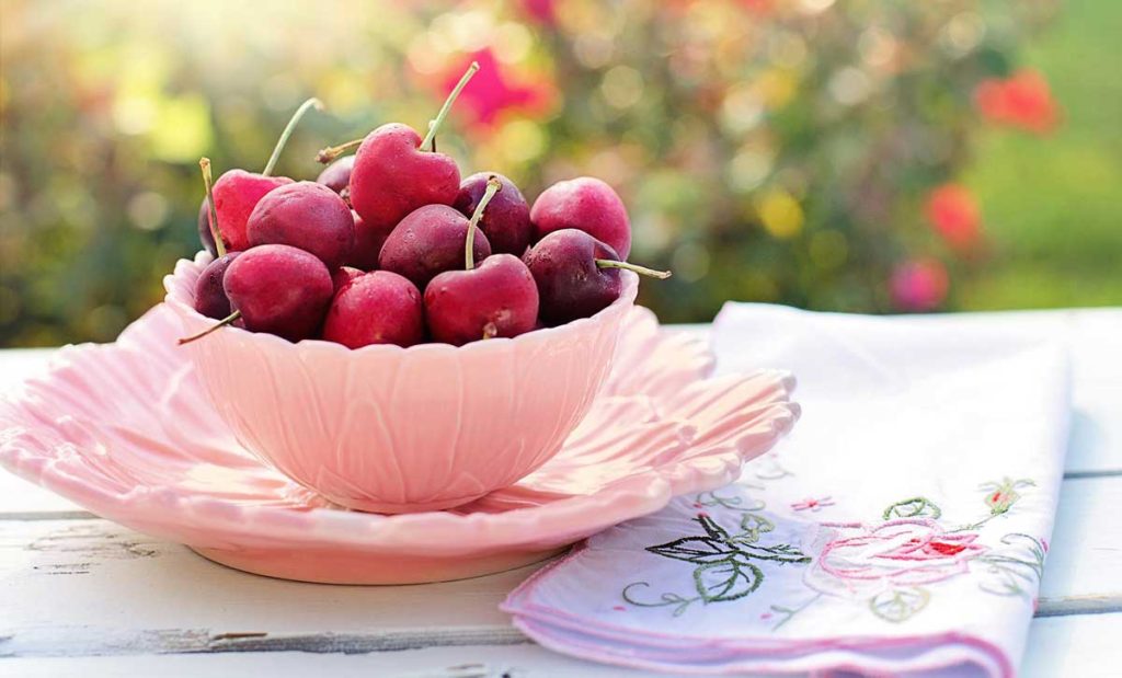 Pink bowl and saucer full of cherries with floral cloth napkin.