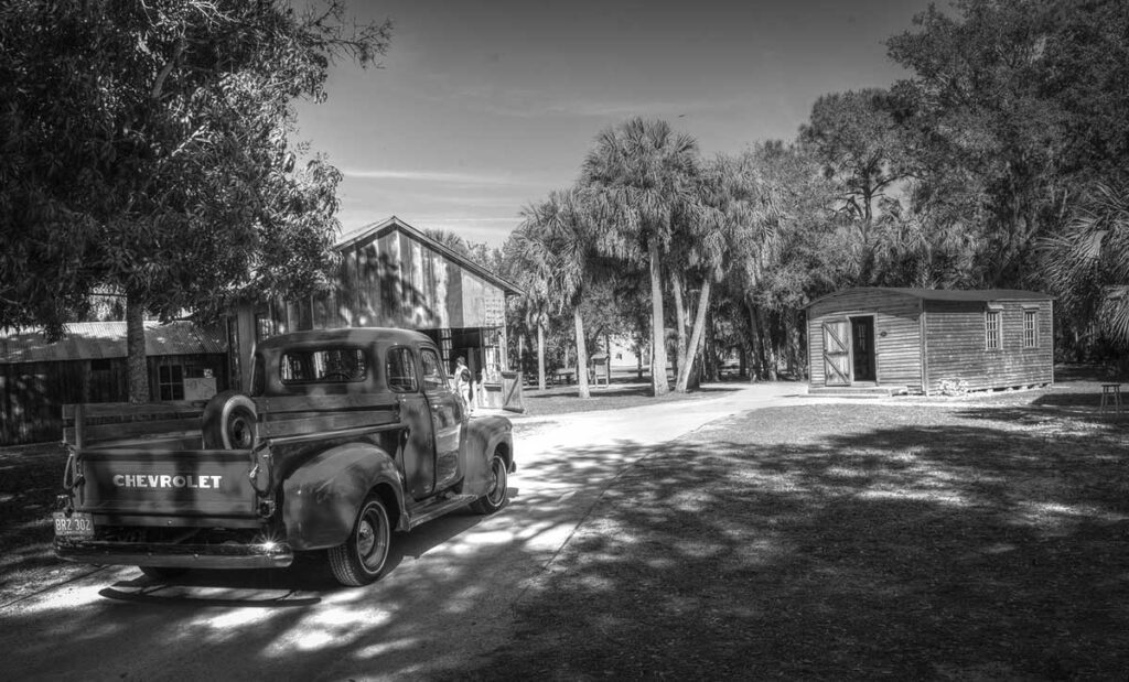 Black and white image of an old chevy truck with old house and outdoor building.