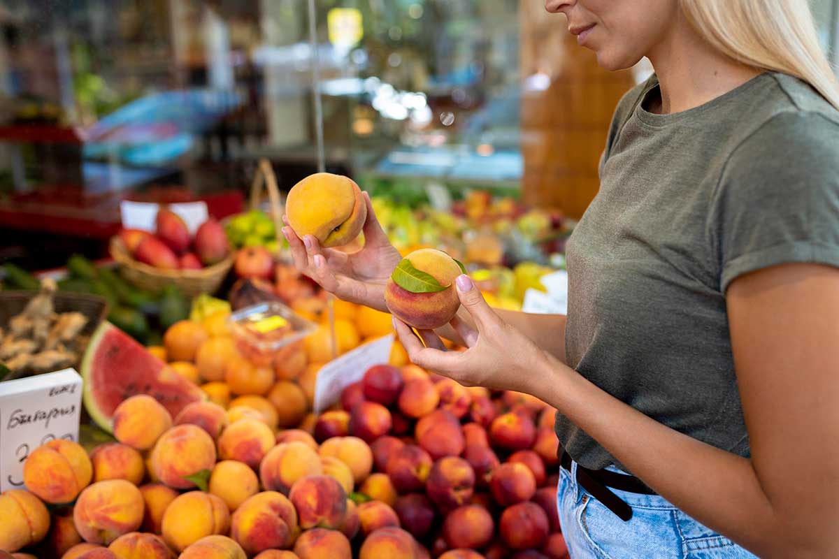 Woman at a fruit and vegetable stand holding a peach i each hand.