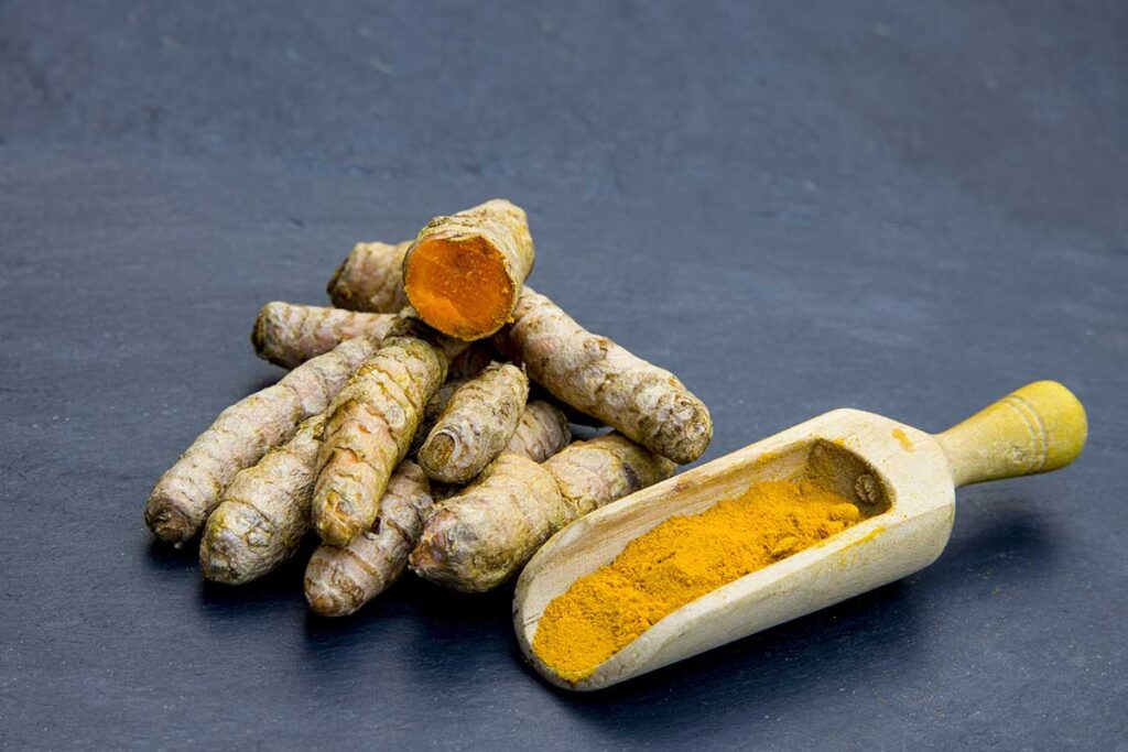 Turmeric root and powder in a wooden scoop on a dark blue background.