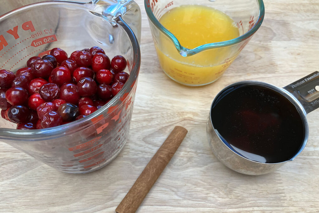 Cranberries and orange juice in two separate Pyrex measuring glasses. Add maple syrup in a measuring cup with a cinnamon stick nearby.