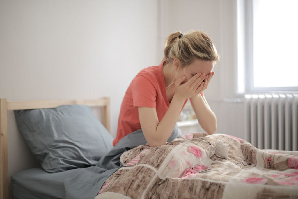 Young blond woman sitting up in bed holding her face.