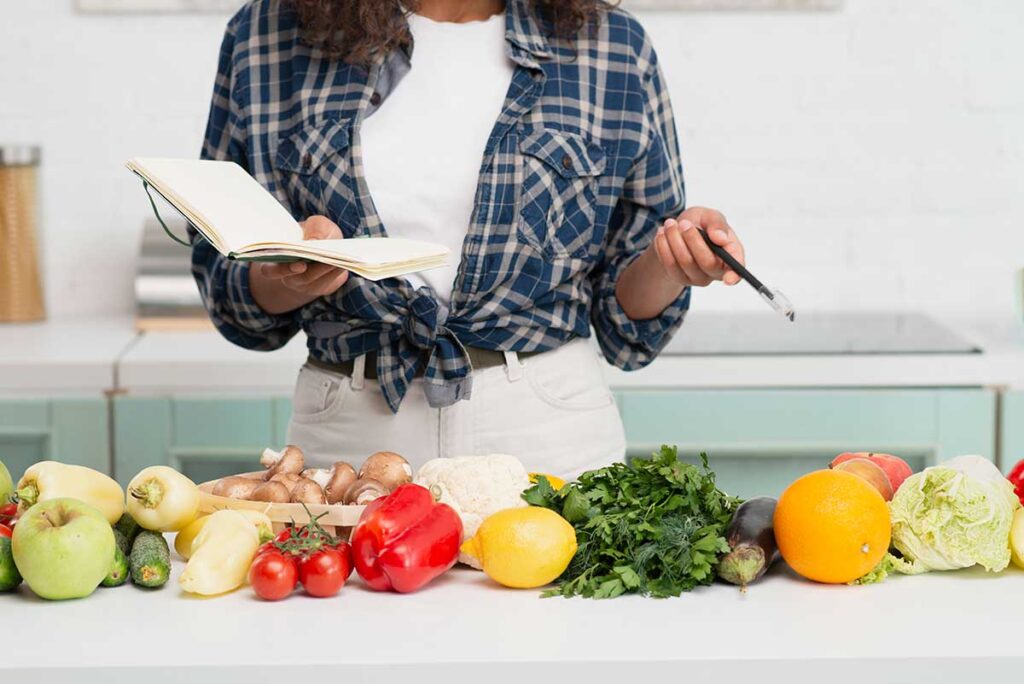 Woman holding notebook looking at vegetables on counter.