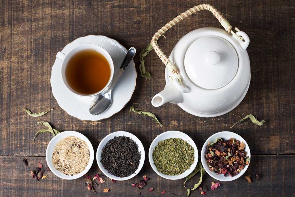 Herbal tea cup and teapot with bowls filled with tea herbs on wooden table.