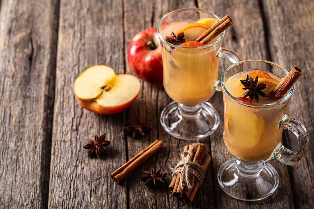 Two glasses of apple drinks with apple slices, cinnamon sticks, and anise on dark wooden table.