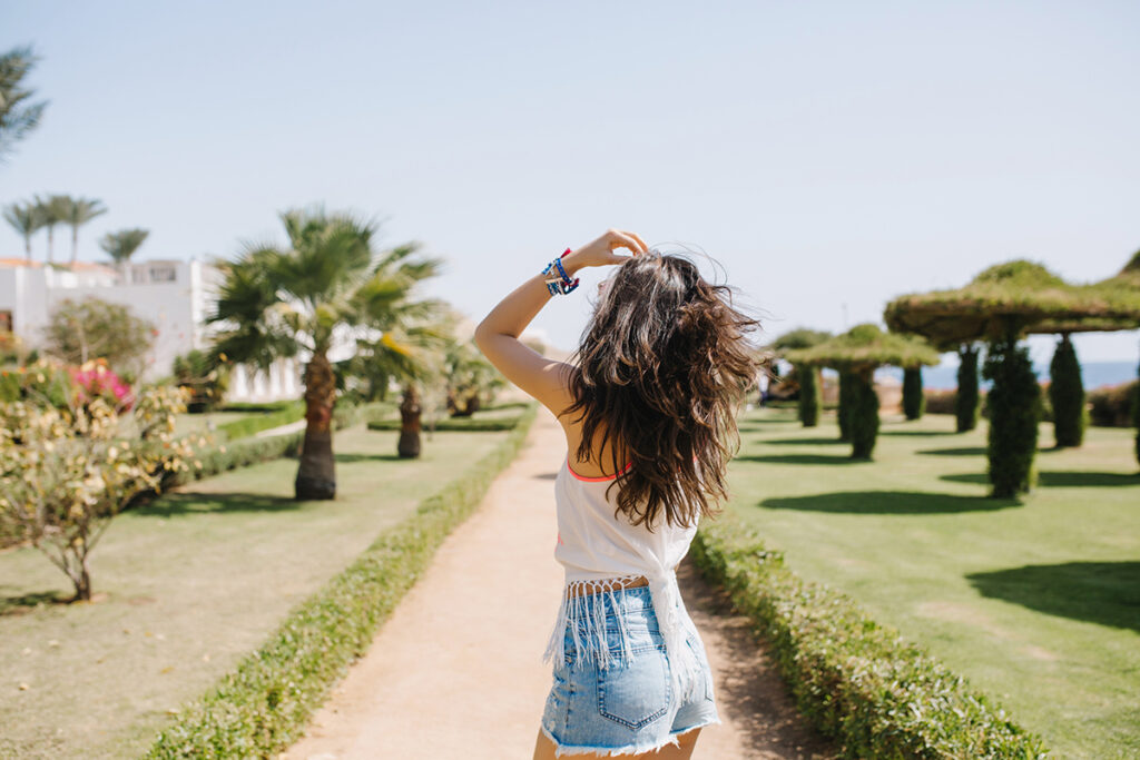 Long-haired woman in white shirt walking on the palm alley under blue sky.