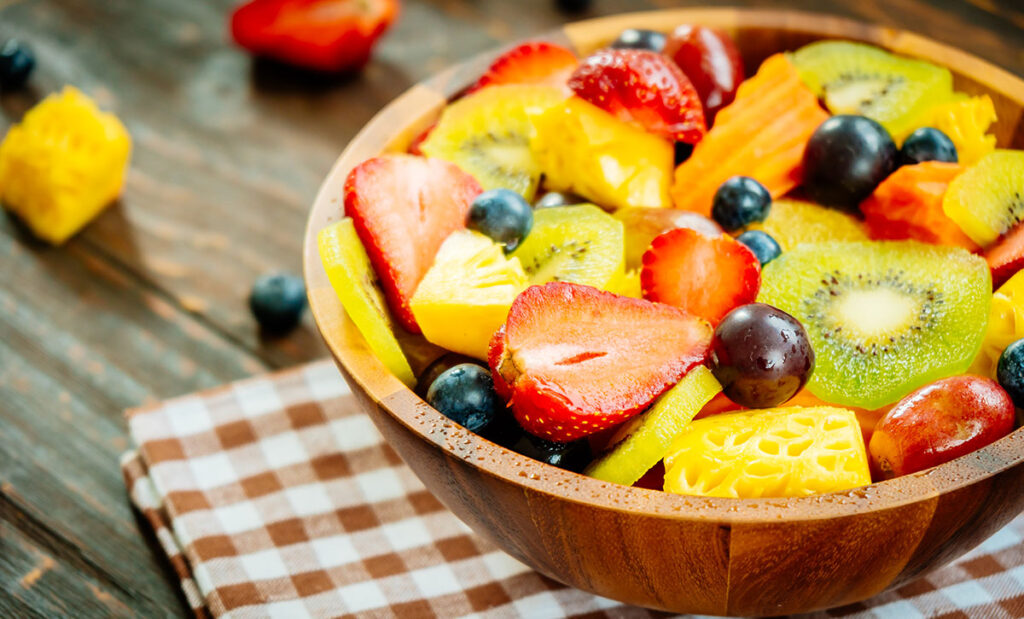 A wooden bowl on a checkered napkin full of assorted fruit.