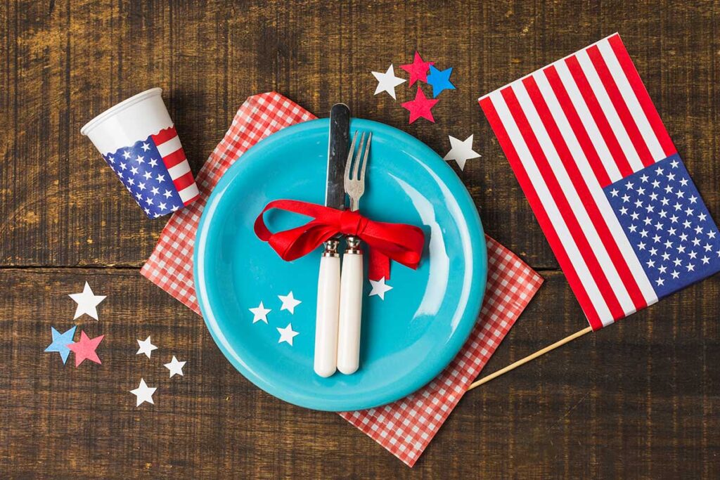 An overhead view of blue plate and cutlery with USA flag and disposable glass on wooden table.