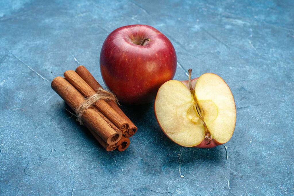 Whole and half red apple with cinnamon sticks tied together on blue background.