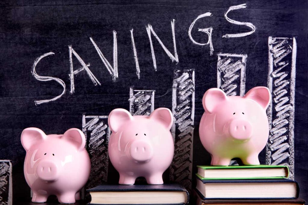 Three pink piggy banks standing on books next to a blackboard with savings chart.