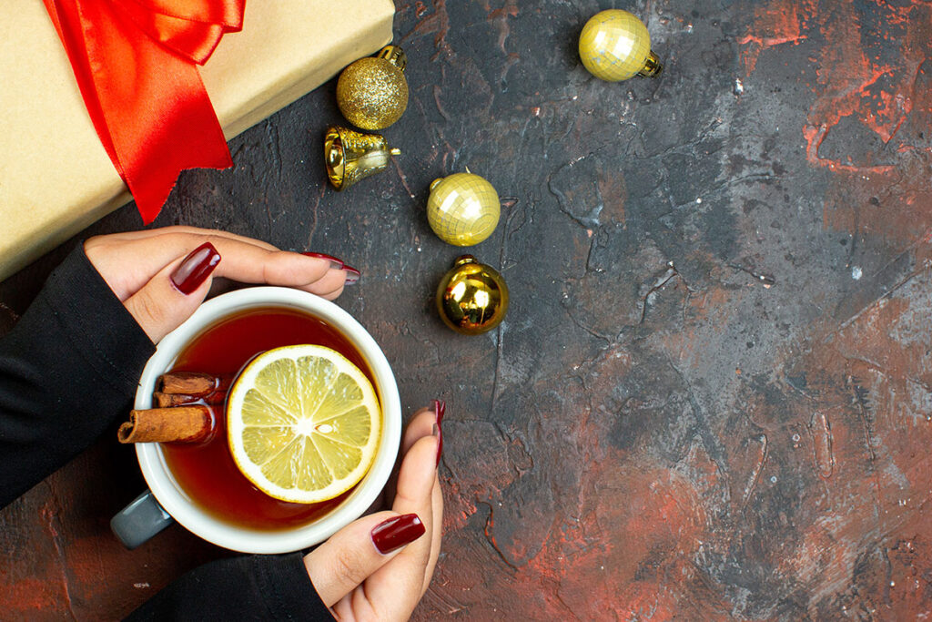 Top view of female hands holding cup of tea flavored by lemon and cinnamon with golden xmas balls on dark red table.