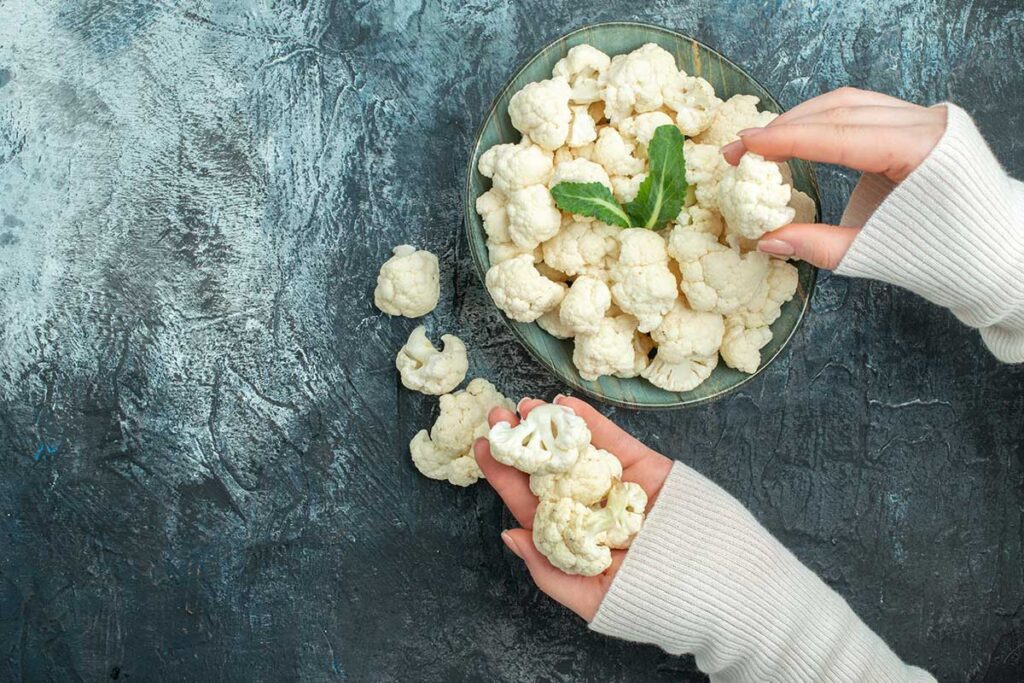 Top view fresh cauliflower inside plate and woman's hands on a light grey table.