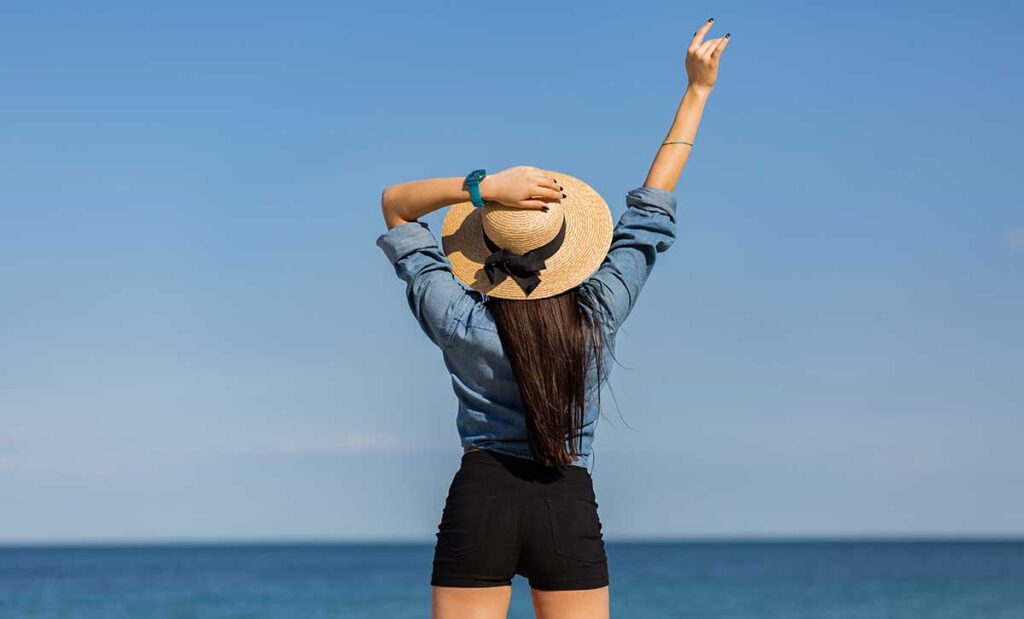 Woman facing ocean holding straw hat with right arm in the air.