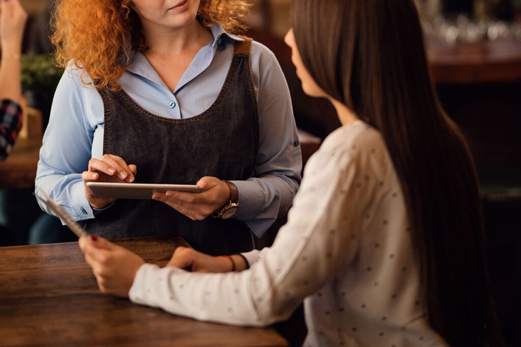 Redhead waitress talking to a woman and writing the order on digital tablet.