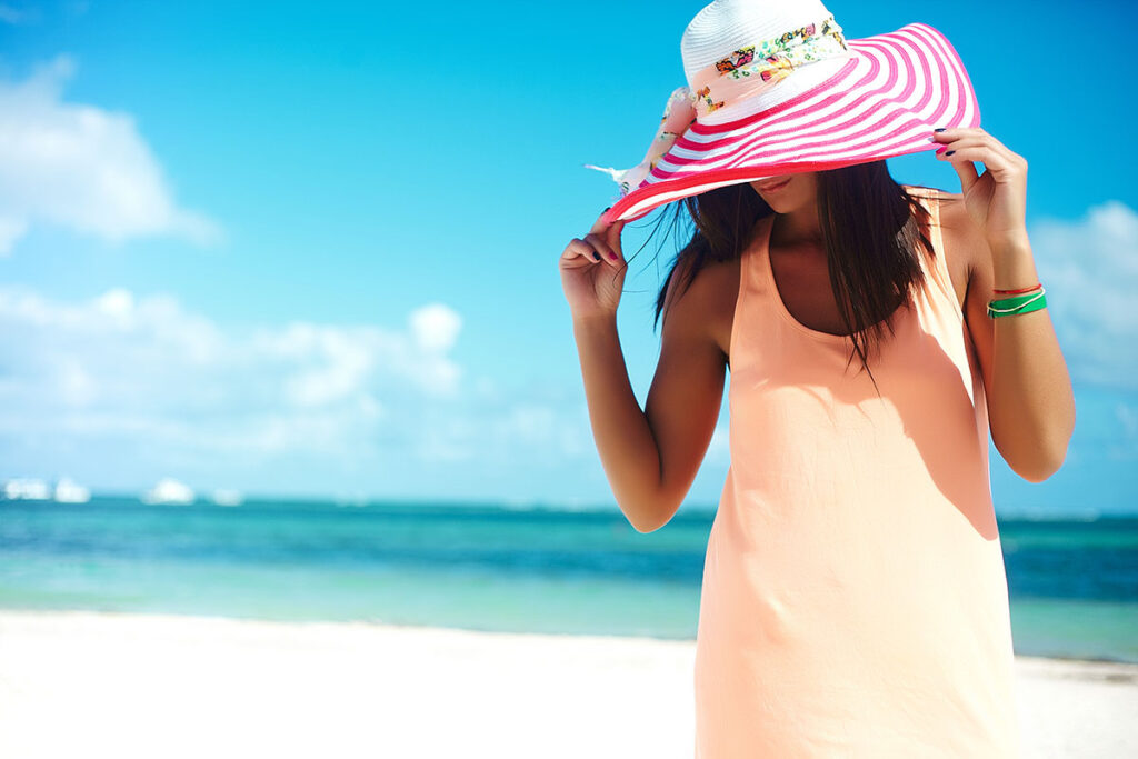 Young woman in colorful sunhat and dress walking near beach ocean on white sand.