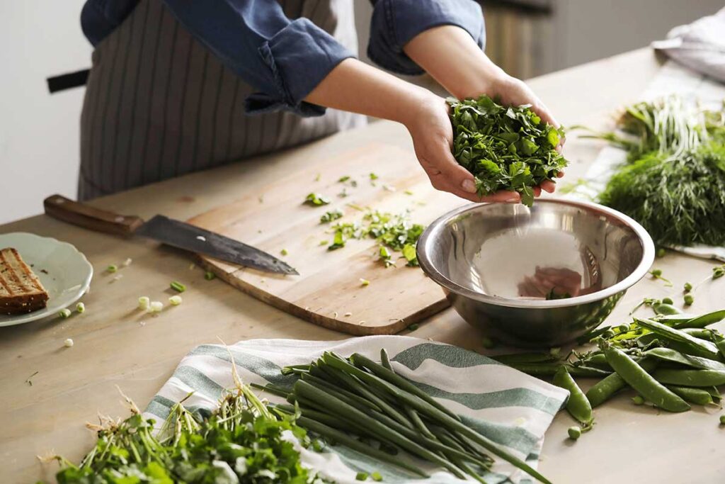 Woman with apron cutting herbs in kitchen and placing them in a bowl.