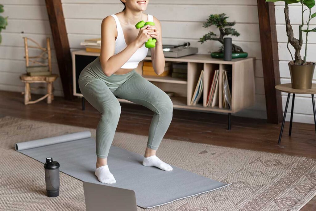 Young woman doing squats at home following fitness workout on laptop with dumbbells at home.