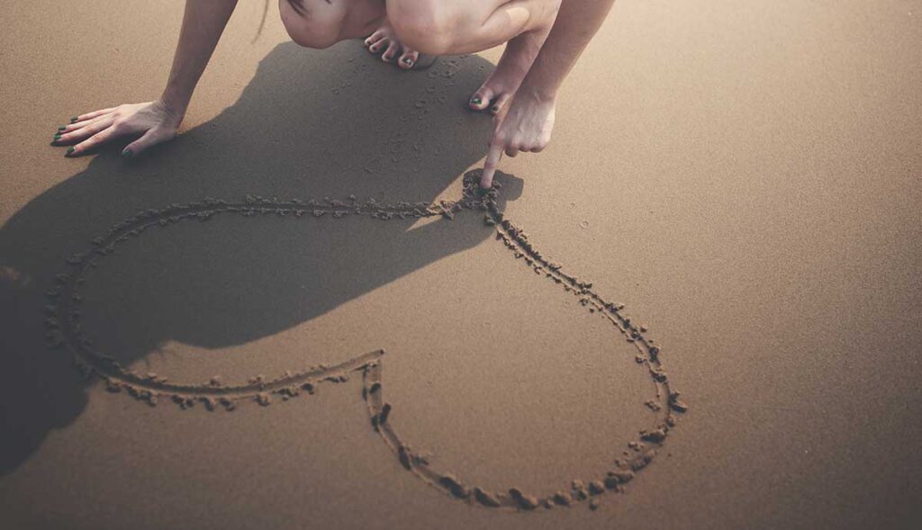 A barefoot woman drawing a heart in the sand.