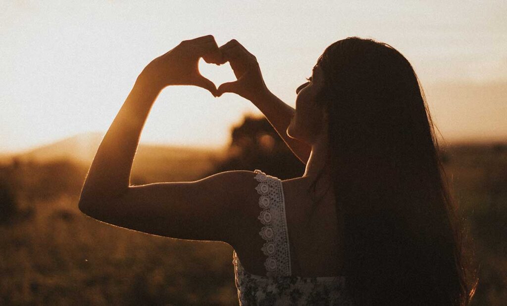 Young woman holding a heart shape with hands during sunset.