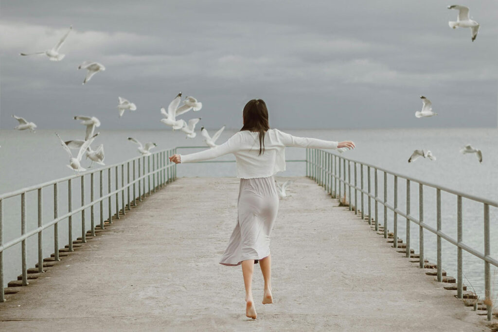 Young barefoot woman on dock and seagulls flying away.