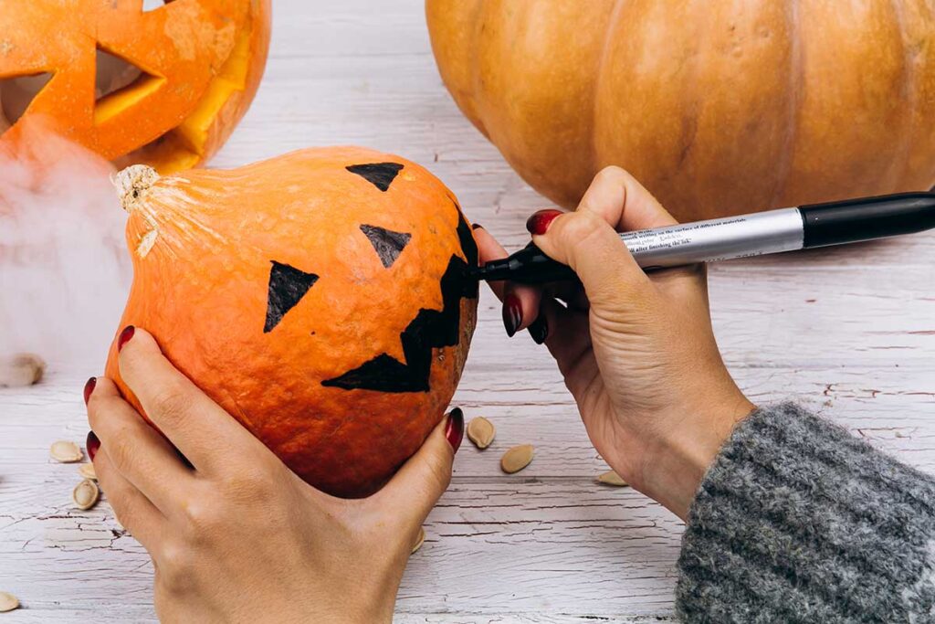 Woman draws a Halloween face on little pumpkin using a black marker.