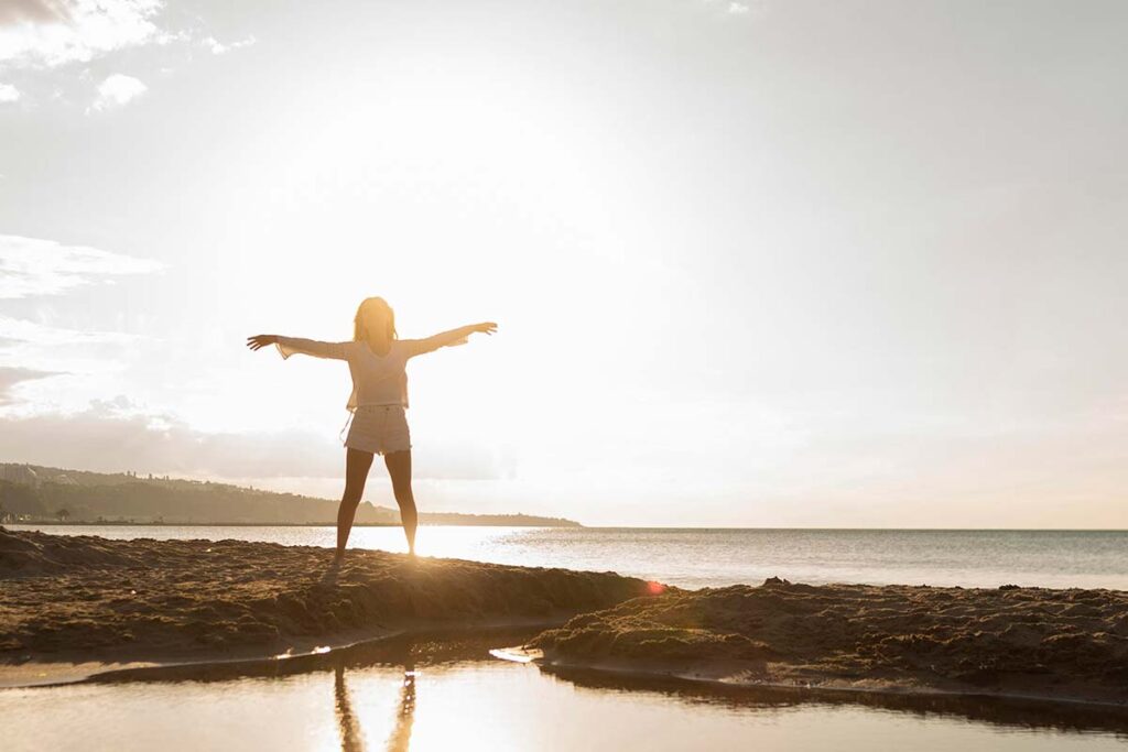 Woman posing with arms stretched out at the beach with sun.