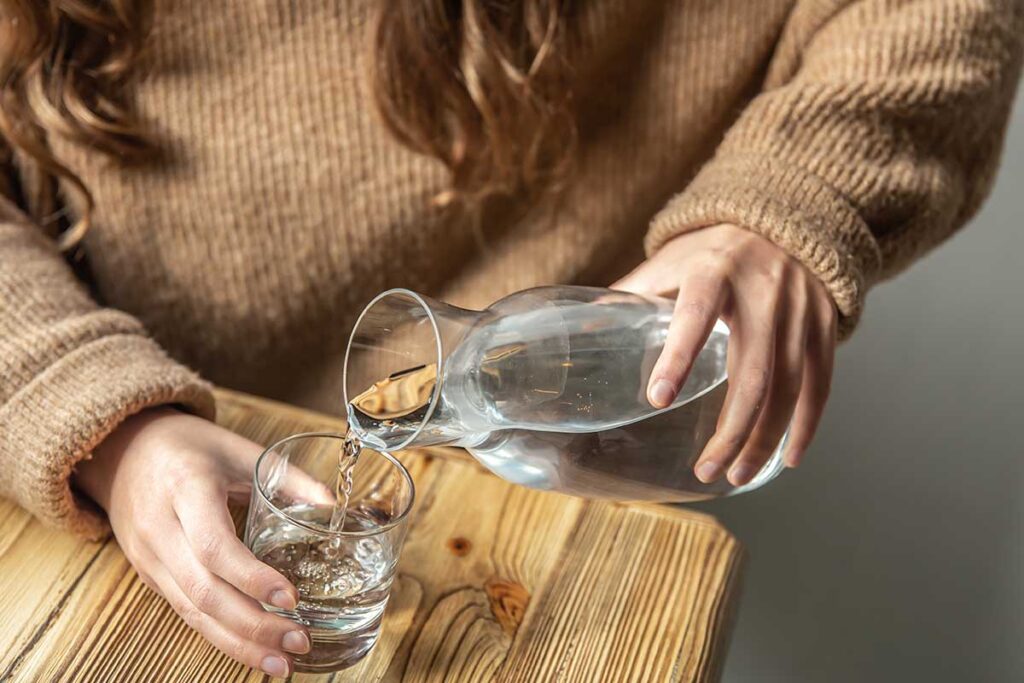 A woman pours water into a glass from a glass decanter.