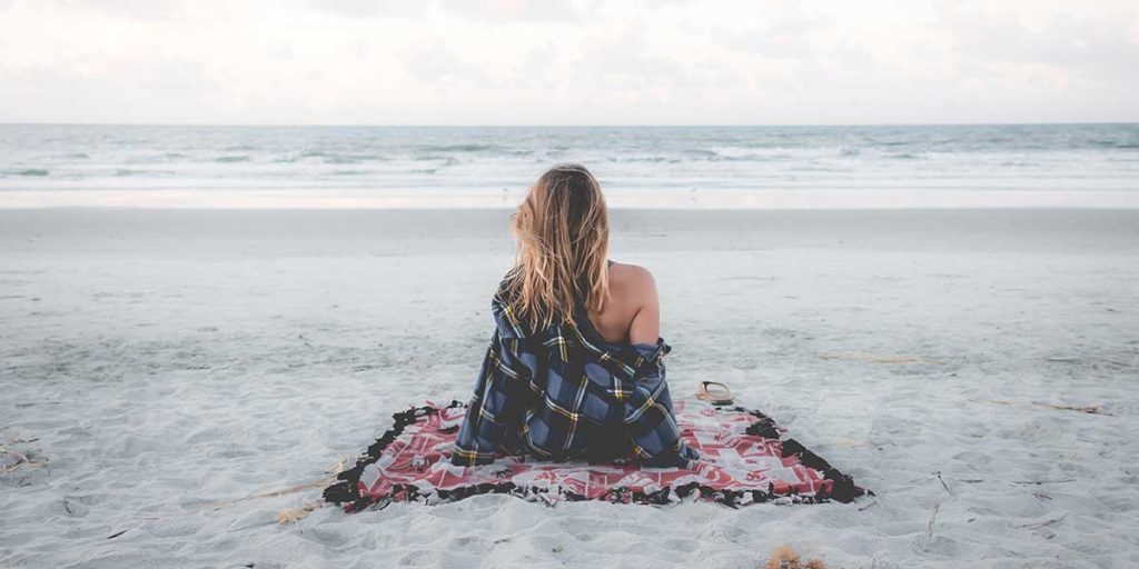 Woman sitting on a blanket on a sandy beach looking at the ocean.