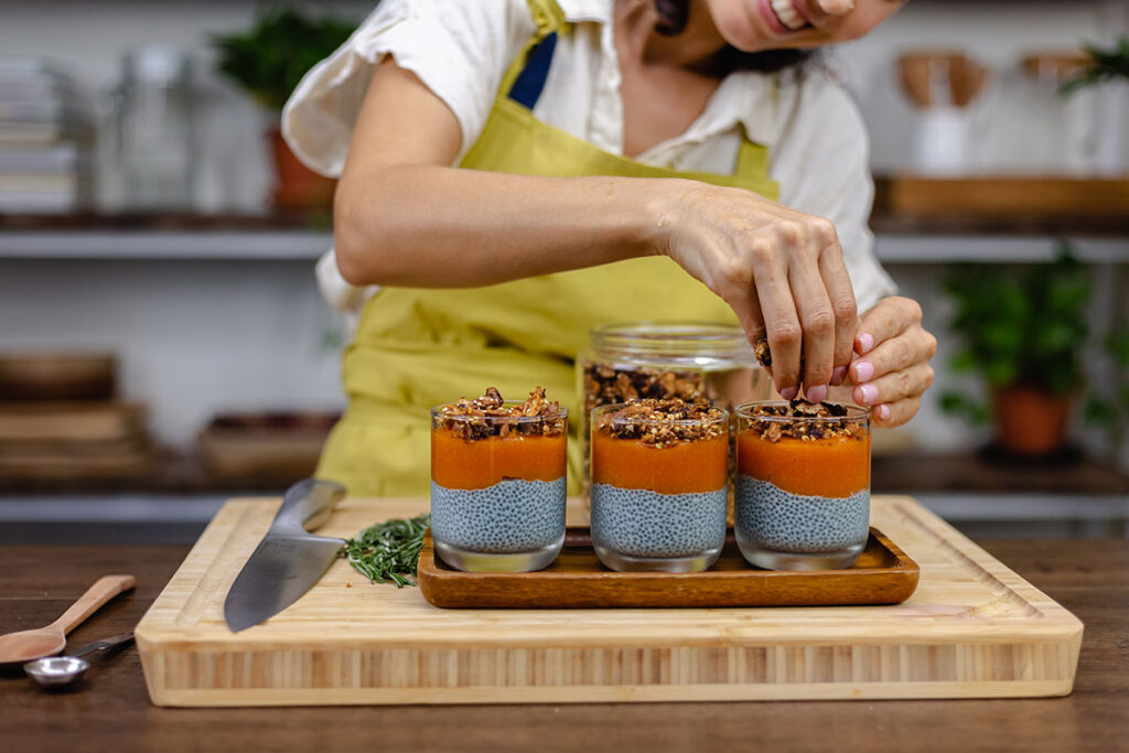 Woman in yellow apron making three jars of chia seed puddings with papaya/mango and homemade granola on cutting board.