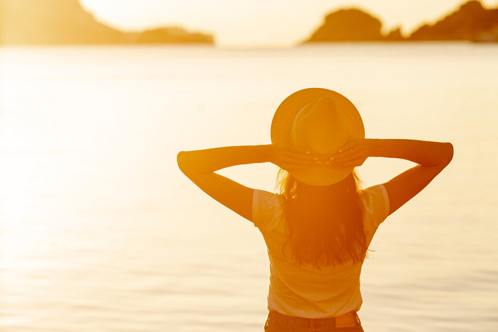 Back of young woman with a hat at sunset on the shore of a lake.