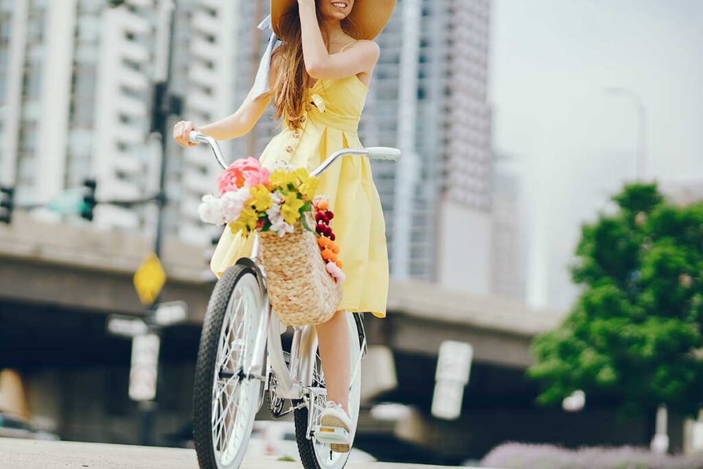 A young woman in a yellow dress riding a bike with flowers in basket.