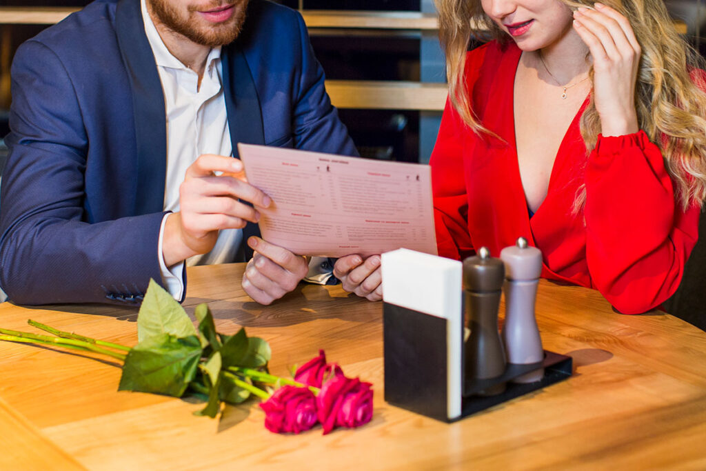 Young blond couple reading menu at table in restaurant and three red roses lying on the table.