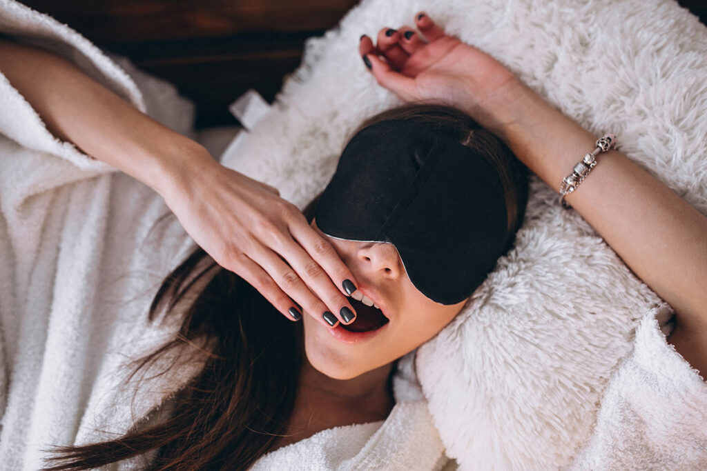 Young woman in bed yawning wearing sleeping mask.