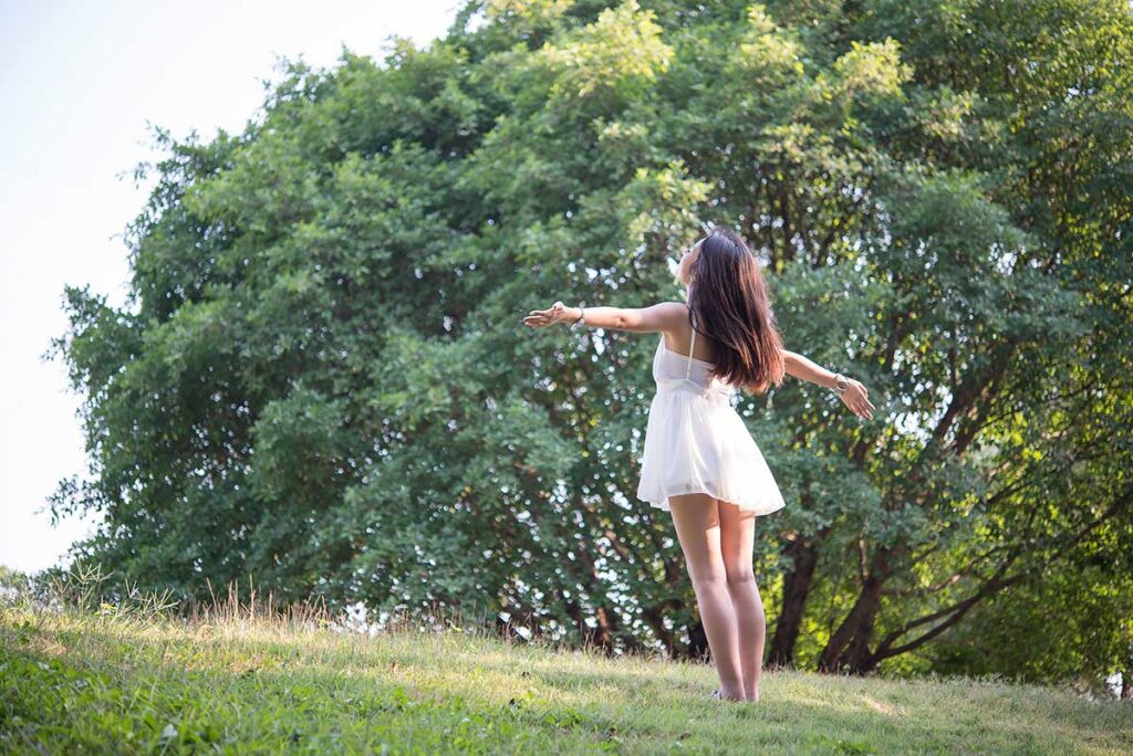 Young barefoot woman in a short white dress on the grass with arms stretched out to each side.