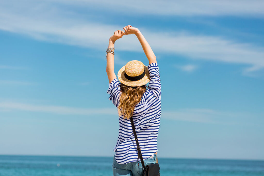 Back of young blonde woman with arms in the air looking at the ocean.