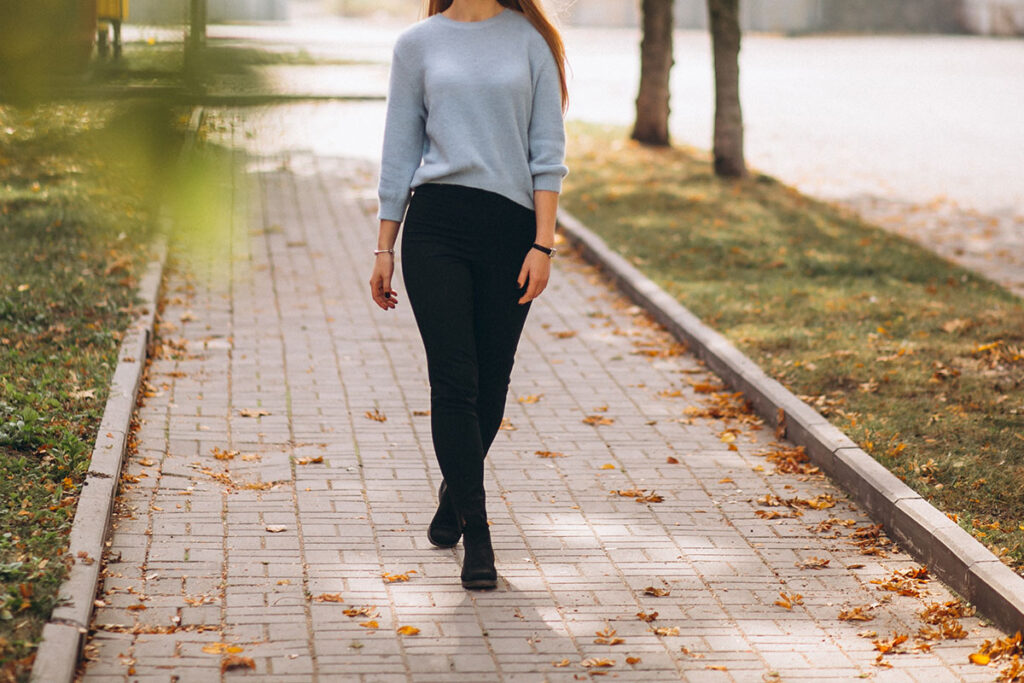 Young woman in black pants and blue sweater walking on brick sidewalk with fallen autumn leaves.